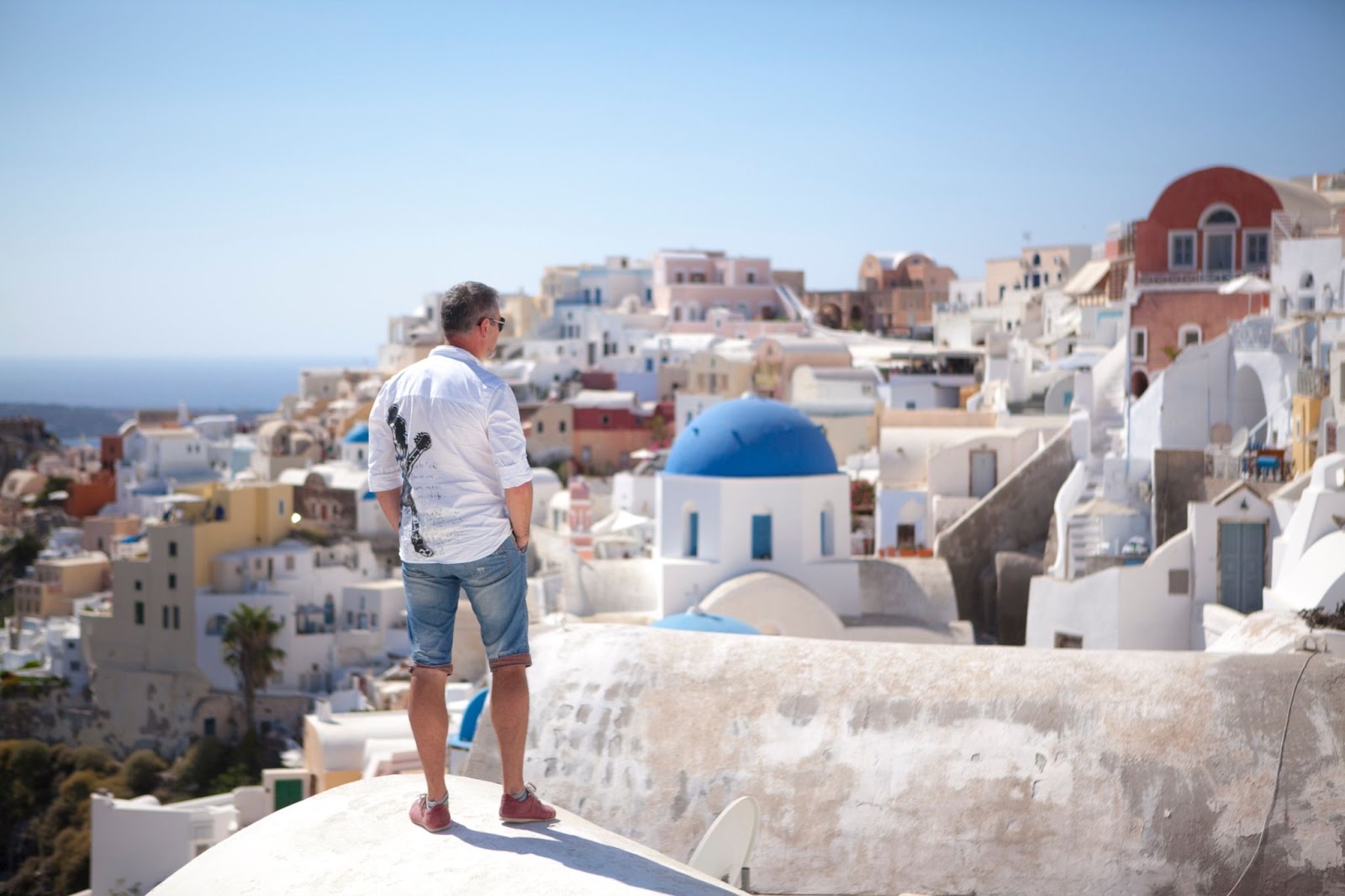 A grown man standing on white semi-circular roof-top classic Greek home. Looking at the panorama of the village on the rocks. Santorini. Greece